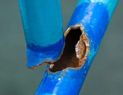 Close-up of a damaged blue pipe showing corrosion and a hole, highlighting risks of burst pipes in winter plumbing systems.