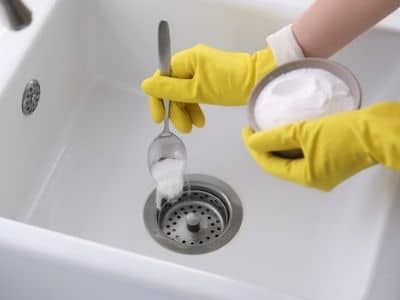 Person wearing yellow gloves pouring baking soda into a kitchen sink drain, demonstrating a method for clearing clogs using baking soda and vinegar.