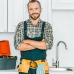 Smiling man in work attire with tool belt, in a clean kitchen representing About Plumbing Inc. plumbing services.