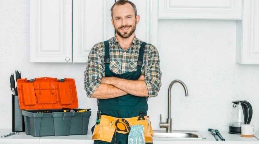 Smiling plumber in a checkered shirt and tool belt, standing confidently in a kitchen with plumbing tools and a sink, representing About Plumbing Inc.'s expert services.