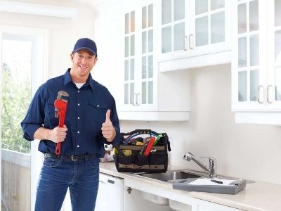 Professional plumber smiling and giving a thumbs up while holding a wrench, standing in a kitchen with tools in a bag, emphasizing plumbing services and expertise.