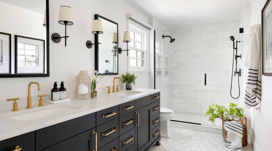 Modern bathroom interior featuring a black vanity with gold fixtures, dual sinks, large mirrors, a glass shower enclosure, and natural light from a window, highlighting stylish design elements for bathroom renovations.