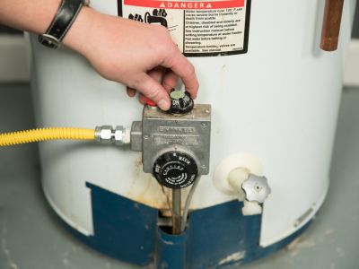 Hand adjusting the control valve on a residential water heater, emphasizing maintenance steps for energy efficiency before leaving home.