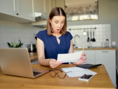 Woman surprised while reviewing high water bill paperwork at kitchen table, emphasizing the impact of water leaks on household expenses.