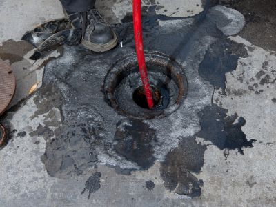 Professional plumber using a hydro jetting hose to clear a clogged drain, with water and debris around the access point, illustrating effective drain cleaning technology.